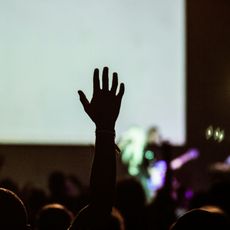 A hand in profile raised above an audience with a lit screen in the background