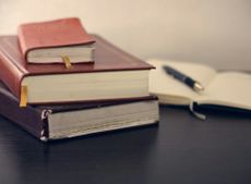 A pile of 3 books with an out-of-focus notebook open in the background with a pen on it